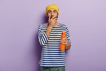 Scared fearful young tourist covers mouth and stares with bugged eyes, wears sailor sweater, prepares for hiking trip, isolated against purple background. Human facial expressions and reaction