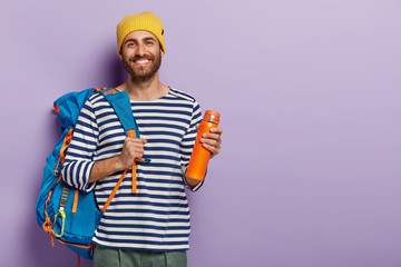 Smiling delighted male tourist has awesome trip, carries big rucksack, drinks coffee from flask,...