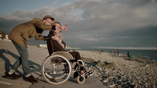 Man is hugging his wife sitting in invalid carriage on sea embankment