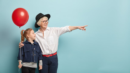 Glad fashionable senior lady embraces grandchild, points into distance over empty blank space, have...