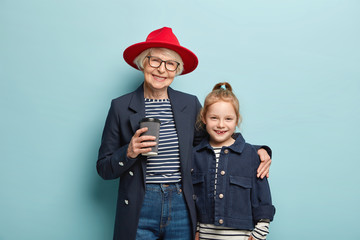 Horizontal view of cheerful grandmother wears red stylish hat, black jacket, drinks takeaway...
