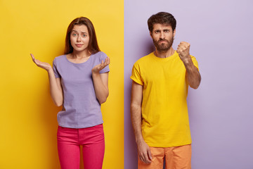 Questioned doubtful woman spreads palms sideways, has unaware facial expression, angry man shows fist at camera, asks not bother him, pose indoor over colorful background. Hesitation and anger
