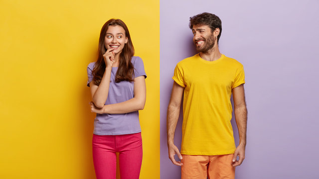 Optimistic young female and male with broad smiles, feel happy, dressed in casual clothes, have fun together, stand against purple and yellow background. People, relations, togetherness concept