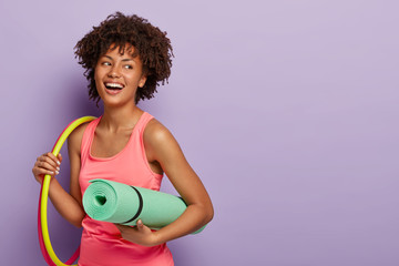 Fitness woman with dark skin, works with hula hoops, holds rolled karemat for training, laughs joyfully, satisfied after workout, looks healthy and fit wears rosy t shirt isolated on purple background - obrazy, fototapety, plakaty