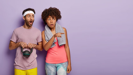 Surprised man and woman go in for sport together, stare with bugged eyes into distance, dressed in sports clothes, hold heavy weight, cannot believe something, blank copy space over purple wall