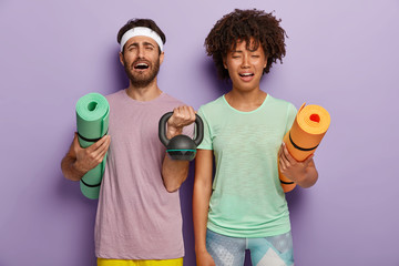 Stressful diverse couple feel tired after sport training with coach, hold fitness mats, weight, dressed in active wear, have miserable facial expressions, isolated on purple background. Bad feelings