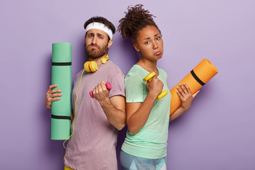 Displeased mixed race couple stand back to each other, raise heavy dumbbells, feel tired afer active sport workout in gym, use karemat, wear casual t shirts, need to take rest, isolated on purple wall