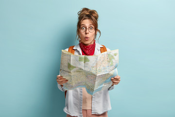 Shocked female European tourist has tour around world, surprised to loose way, reads map, wears round spectacles, carries backapck, poses over blue background. Summer travel concept. Sightseeing
