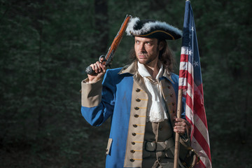 Portrait of man dressed as soldier of War of Independence United States aims from pistol with flag....