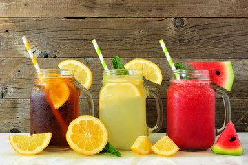 Variety of summer drinks in mason jar glasses with fruit against a dark wood background. Iced tea, lemonade and watermelon juice.