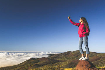 Teenager standing on a mountain cliff edge gorgeous view to forest sea and clouds, taking selfie with smartphone. Generation z are always connected Adventure freedom tourism carefree and youth concept