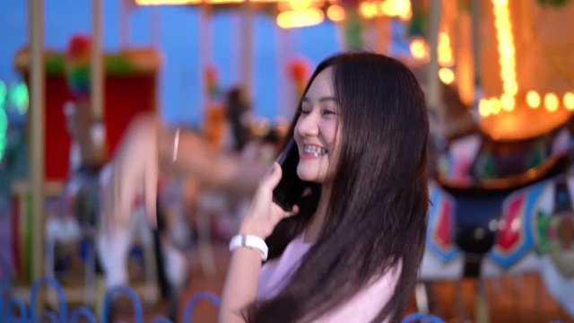 Asian beautiful woman call a friend with smartphone at amusement park carnival. Woman happy together friendship travel night funfair festival 4k resolution.