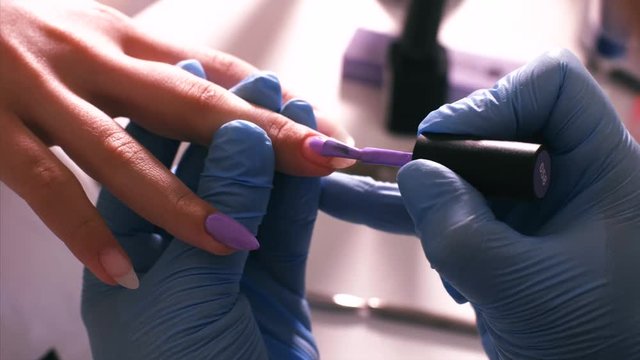 Manicurist master applies a violet coat of shellac using brush on client's nails, hands in gloves closeup. Painting nails in beauty salon.
