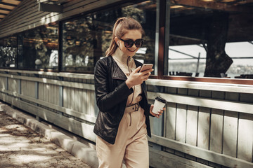 Portrait of Stylish Girl Wears Women Overalls and Black Leather Jacket Drink Coffee, Using Smartphone