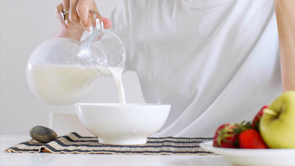 Portrait of woman in white t-shirt is preparing for eat cornflakes with milk and fruits for breakfast. She poured milk in a bowl while sitting at table on white background, hands close-up.