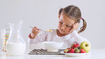 Portrait of sad small girl doesn't want to eat cornflakes with milk for breakfast. She is looking...