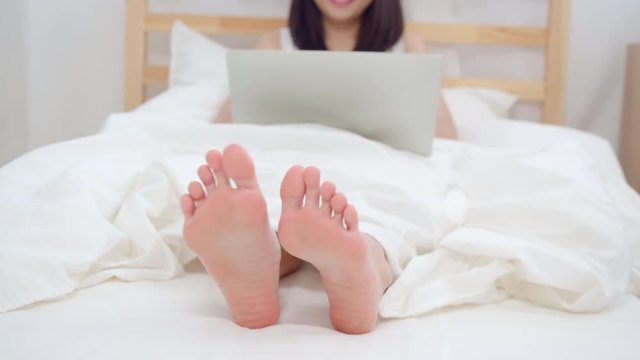 Young business freelance Asian woman working on laptop checking social media while lying on the bed when relax in bedroom at home. Lifestyle women at house concept.