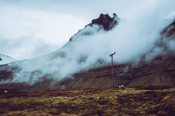 Fototapeta premium Blue and purple colors of peaceful Icelandic scenery. Dark green grass, electric grids, and dreamlike clouds covering sharp mountain range.