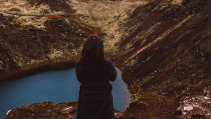 Brunette girl in the gray coat standing on the edge of the red rock surrounding the lake of Kerid (Kerith) located in a volcanic crater. Dreaming and traveling concept. Look from behind.