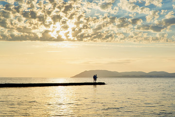 Silhouette of two people on the pier fishing in the sea at sunset against the mountains