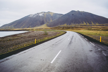 Typical dark gray wet asphalt of beautiful Icelandic roads. Highway by the lake and bold mountains covered by gray clouds. Pale green grass.