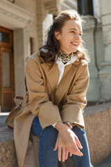 Young beautiful girl with long hair in beige coat on a sunny day sitting on a curb and smiling. Street style portrait