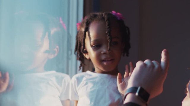 Adorable curious little African american girl touching a man's hand in sunlight near the window. Portrait of lovely african kid with braids at home.