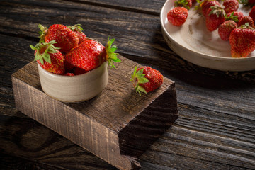 Ripe strawberries in a cup and on a ceramic plate lie on a wooden background.