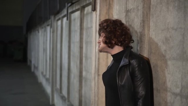 Young caucasian funny guy actor dressed like woman, wearing black leather jacket with blouser and put on head brown wig, sings outside standing next to beige wall in street in the evening.