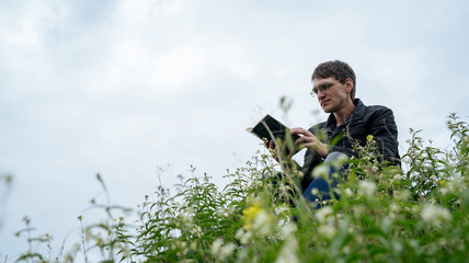 Side view of young handsome man in glasses and warm jacket reading book in open space on background of green hills