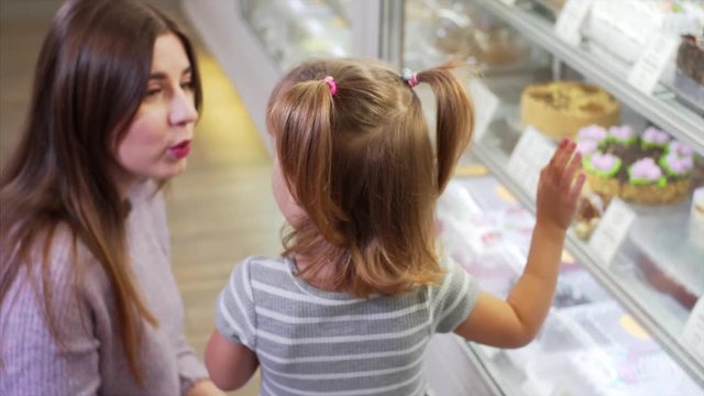 Cute girl and her mom choose delicious cakes for dessert in coffee shop