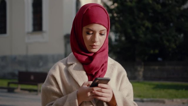 Sociable arab girl using a smartphone on the street at sunlight. Portrait of pretty Muslim woman in red hijab smiling at camera.