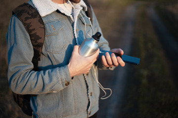 Close-up of hands of young modern guy with backpack, holding the coffee cup and using smartphone with earphones
