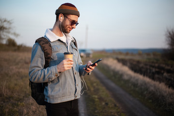 Portrait of young guy holding cup of hot coffee and smartphone with earphones outdoor