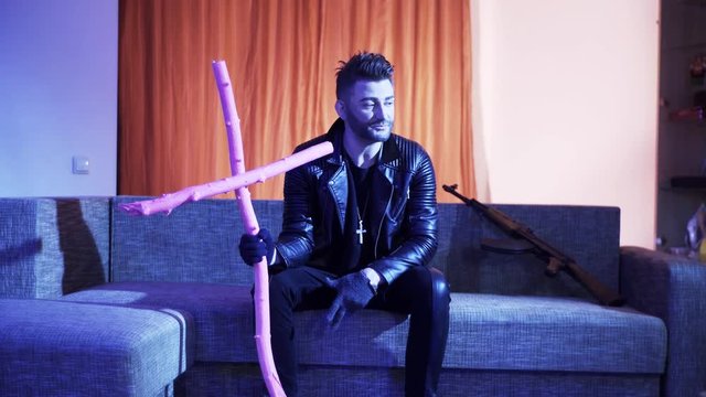 Attractive guy with stylish haircut, wearing black leather jacket, gold cross with chain and gloves, sits on modern grey sofa with wooden pink cross and machine gun, sings and looks at camera.
