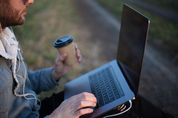 Close-up of young sitting guy with laptop on legs and cup of coffee in hand