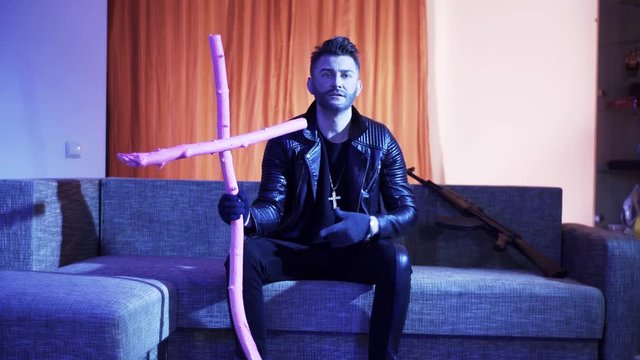 Pretty bearded guiy with nice haircut, wearing black leather jacket, gold cross with chain and gloves, sits on modern grey sofa with wooden pink cross and machine gun, sings and looks at camera.