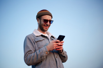 Portrait of smiling modern guy with smartphone in hands on background of blue sky
