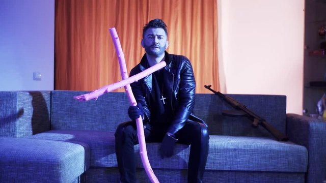 Good-looking young bearded man wearing black leather jacket, gold cross with chain and gloves, sits on modern grey sofa with wooden pink cross and machine gun, sings and looks at camera.