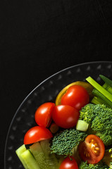 Vegetable salad. Broccoli, tomatoes, cucumbers, green onions on a black plate on a black wooden background.  Place the text at the top left. The view from the top.