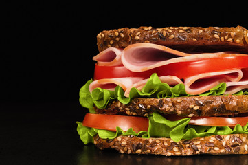 Homemade big sandwich of toasted grain bread with ham, tomato, vegetation on a black table on a black background. Close up. Left place under the text.