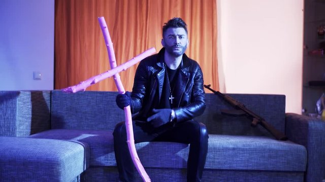 Sexy looking bearded man with stylish haircut, wearing leather jacket, gold cross with chain and gloves, sits on modern grey sofa with wooden pink cross and machine gun, sings and looks at camera.