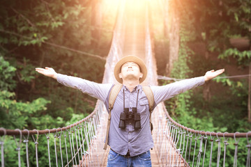 Happy celebrating winning success male tourist standing elated with arms raised up above on pedestrian hanging bridge over river in tropical forest