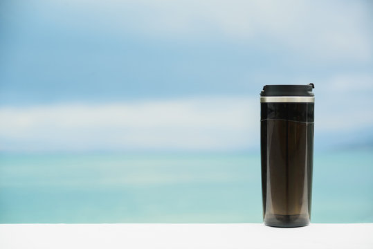 plastic black thermos mug without handle on blurred sea and sky background with copy space, zero waste and my cup please concept, still life mockup or template, horizontal stock photo image