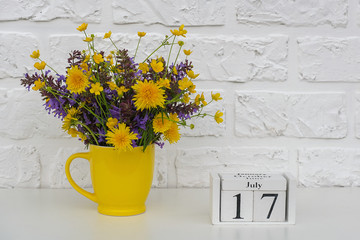Wooden cubes calendar July 17 and yellow cup with bright colored flowers against white brick wall. Template calendar date for your design Copy space