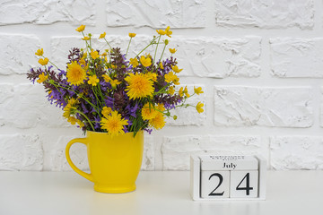 Wooden cubes calendar July 24 and yellow cup with bright colored flowers against white brick wall. Template calendar date for your design Copy space.