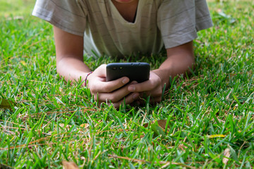 Asian teen boy lying on grasses ground in the garden and using smartphone happily.
