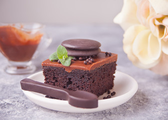 A piece of homemade chocolate cake on the plate with icing, mint leaf and chocolate spoon on the gray table