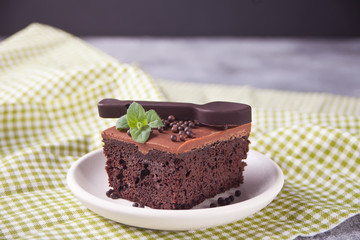 A piece of homemade chocolate cake on the plate with icing, mint leaf and chocolate spoon on the gray table