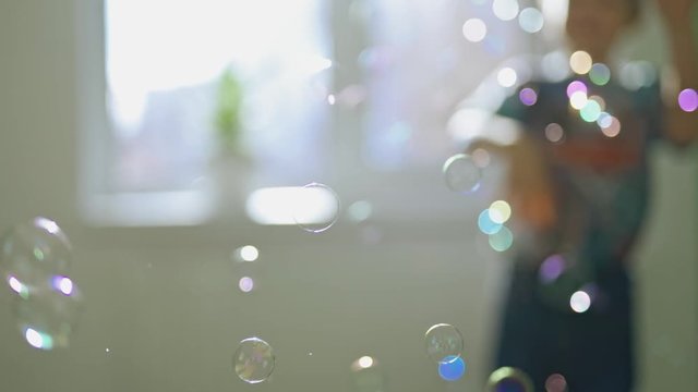 Round colorful bubbles flying in the room on the blurred background with a boy. Beautiful soap bubbles and a silhouette of a boy playing with them indoors.